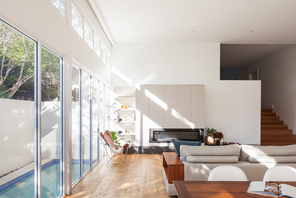 Cette image montre une salle de séjour minimaliste ouverte et de taille moyenne avec un mur blanc, un sol en bois brun, une cheminée ribbon, un sol marron et un manteau de cheminée en bois.