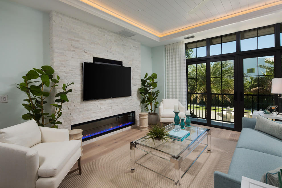 Modelo de sala de estar costera con paredes azules, suelo de madera clara, chimenea lineal, marco de chimenea de piedra, televisor colgado en la pared y suelo beige