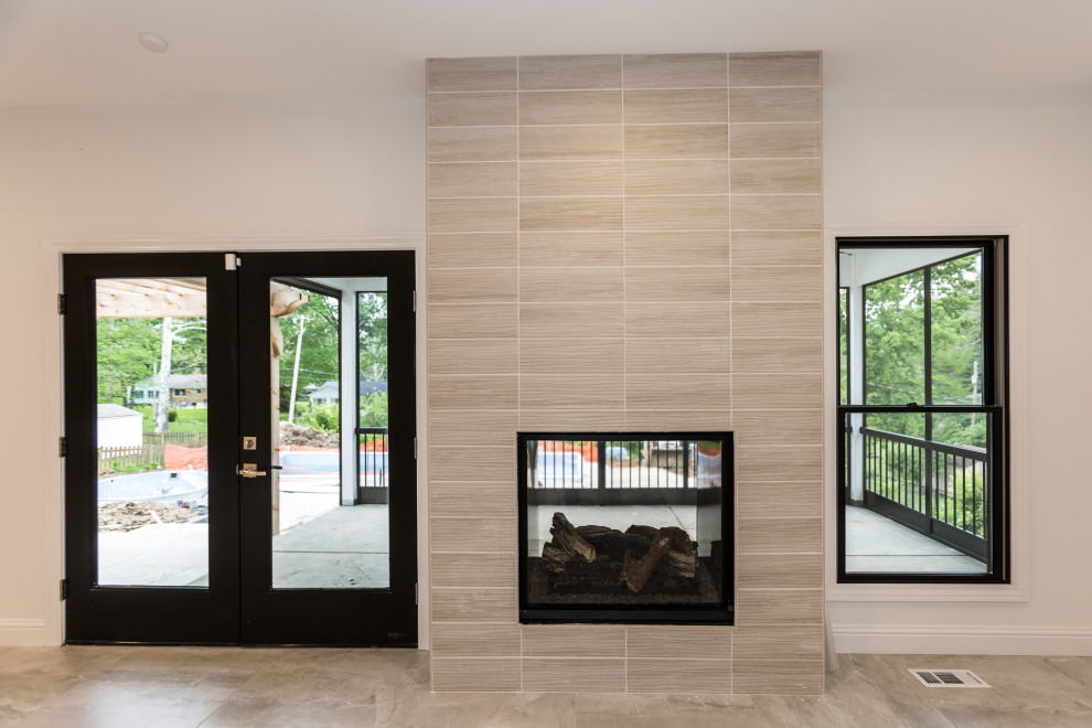 Пример оригинального дизайна: большая открытая гостиная комната в стиле неоклассика (современная классика) с белыми стенами, полом из керамической плитки, двусторонним камином, фасадом камина из плитки, телевизором на стене и серым полом