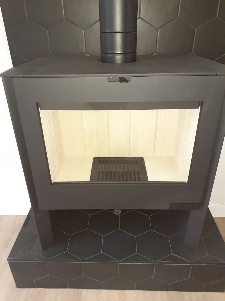 This is an example of a small industrial open plan games room with a freestanding tv, black walls, ceramic flooring, a wood burning stove, a tiled fireplace surround and black floors.