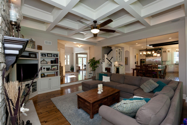 Cette photo montre une grande salle de séjour craftsman ouverte avec un mur gris, un sol en bois brun, une cheminée standard, un manteau de cheminée en pierre, un téléviseur d'angle et un sol marron.