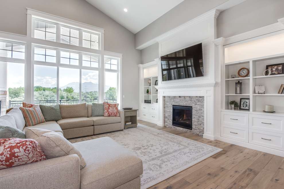Ejemplo de sala de estar abierta de estilo americano de tamaño medio con paredes grises, suelo de madera clara, todas las chimeneas, marco de chimenea de piedra y televisor colgado en la pared