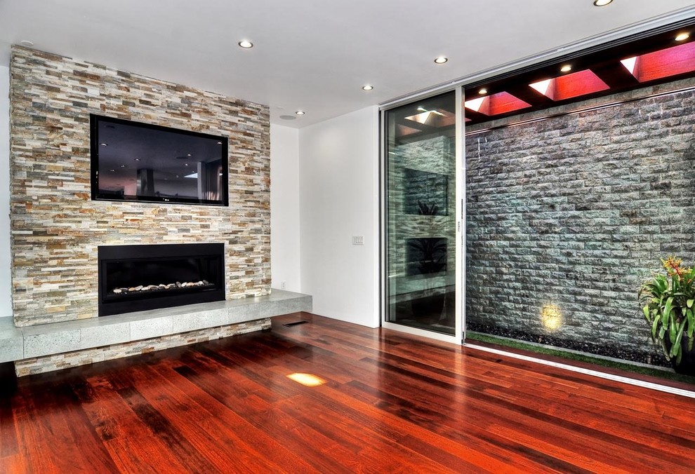 Modelo de sala de estar abierta contemporánea grande con paredes blancas, suelo de madera oscura, chimenea lineal, marco de chimenea de piedra y televisor retractable