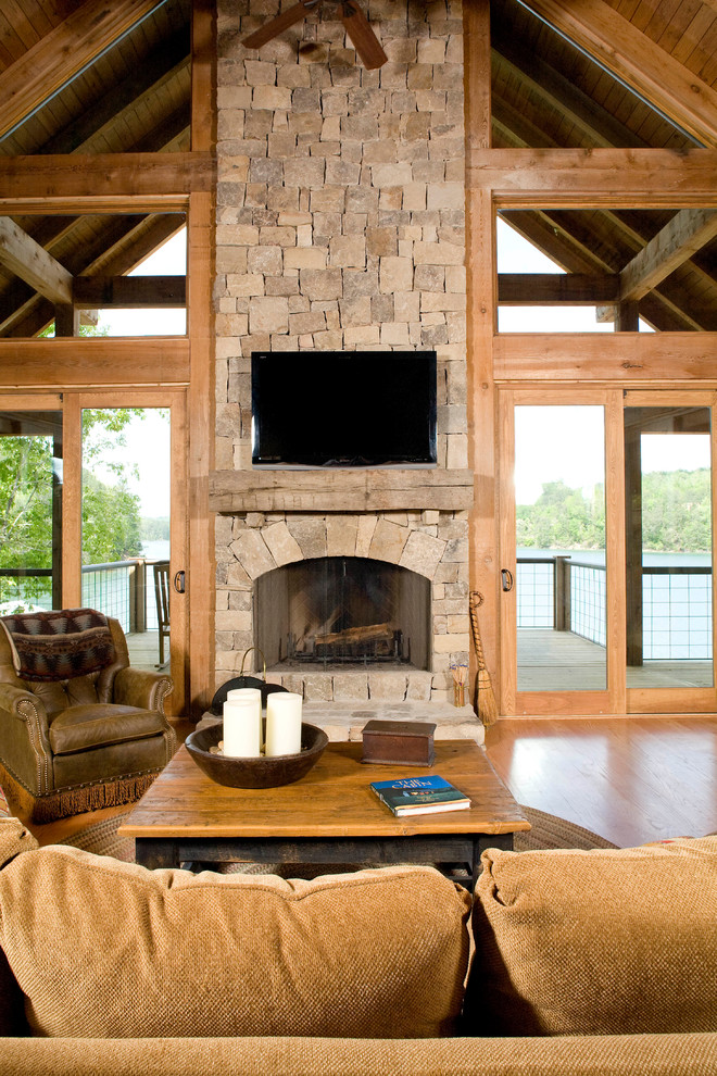 Cette image montre une grande salle de séjour craftsman ouverte avec parquet clair, une cheminée standard, un manteau de cheminée en pierre, un téléviseur fixé au mur, un mur marron et un sol marron.