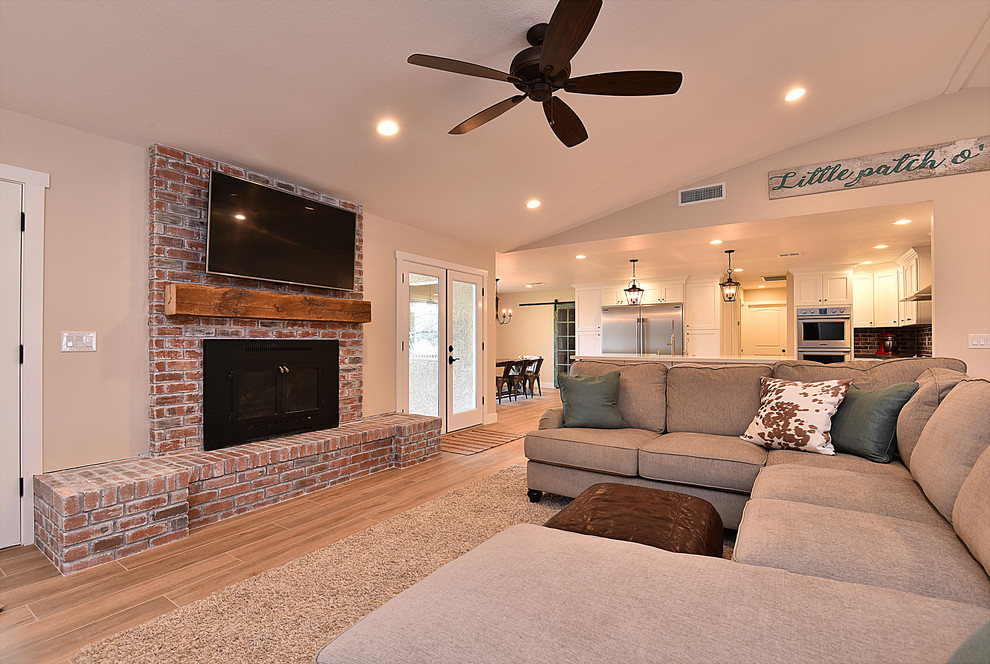 This is an example of a large country open plan games room in Phoenix with beige walls, light hardwood flooring, a standard fireplace, a brick fireplace surround, a wall mounted tv and brown floors.