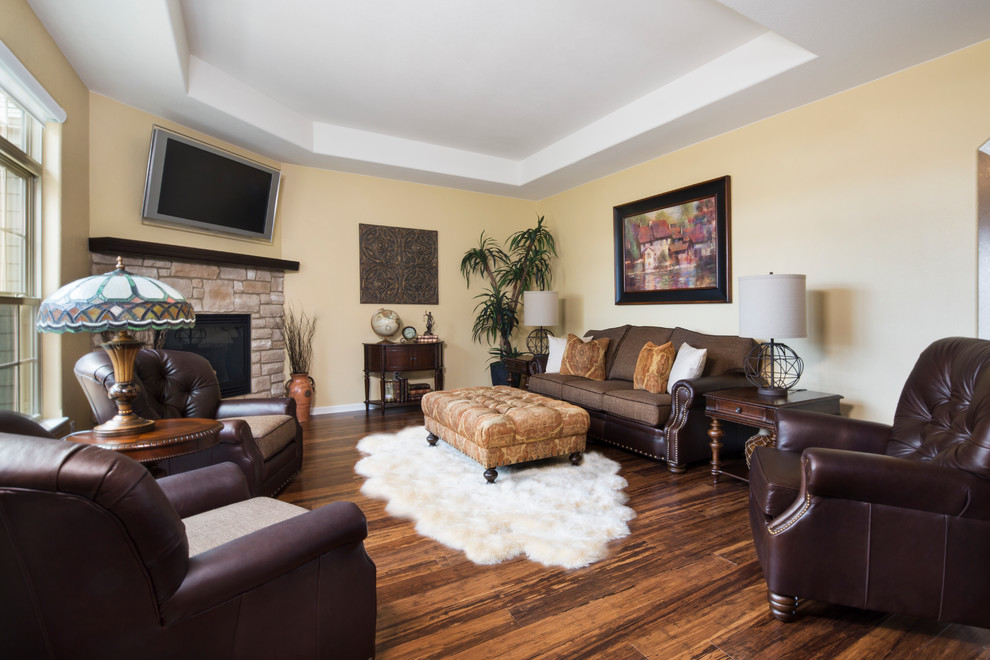 Imagen de sala de estar cerrada clásica grande con paredes amarillas, suelo de madera en tonos medios, chimenea de esquina, marco de chimenea de piedra y televisor colgado en la pared