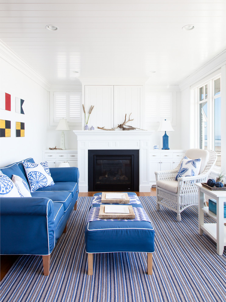 На фото: гостиная комната в морском стиле с белыми стенами, паркетным полом среднего тона, стандартным камином, коричневым полом, скрытым телевизором и синим диваном с