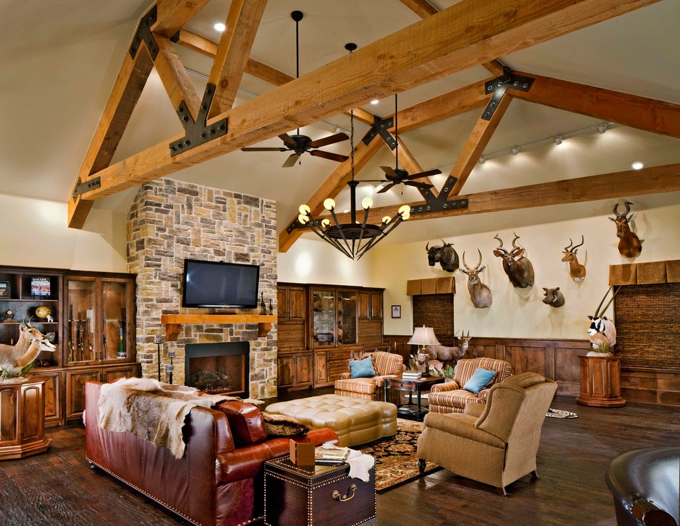 Imagen de sala de estar rural con paredes beige, suelo de madera oscura, todas las chimeneas, televisor colgado en la pared y marco de chimenea de piedra