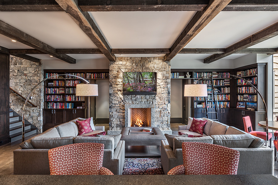 Exemple d'une grande salle de séjour montagne ouverte avec une bibliothèque ou un coin lecture, un mur gris, sol en béton ciré, une cheminée standard, un manteau de cheminée en pierre, un téléviseur fixé au mur et un sol gris.
