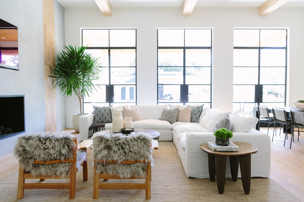Modelo de sala de estar abierta costera con paredes blancas, suelo de madera clara, chimenea lineal, televisor colgado en la pared y suelo beige