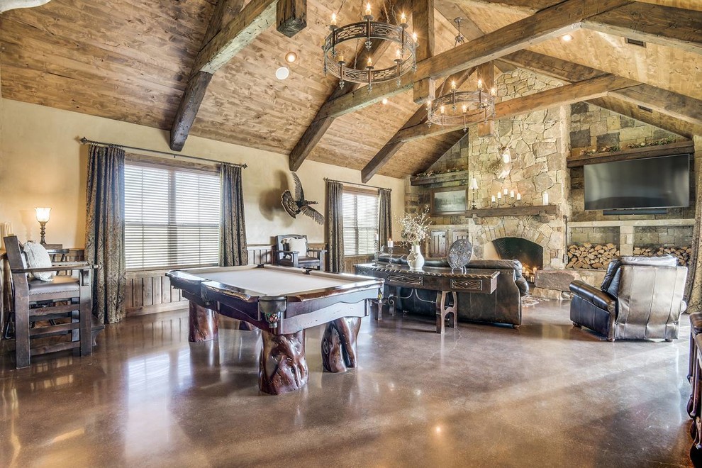 This is an example of a large rustic open plan games room in Austin with a game room, beige walls, concrete flooring, a standard fireplace, a stone fireplace surround, a wall mounted tv and brown floors.