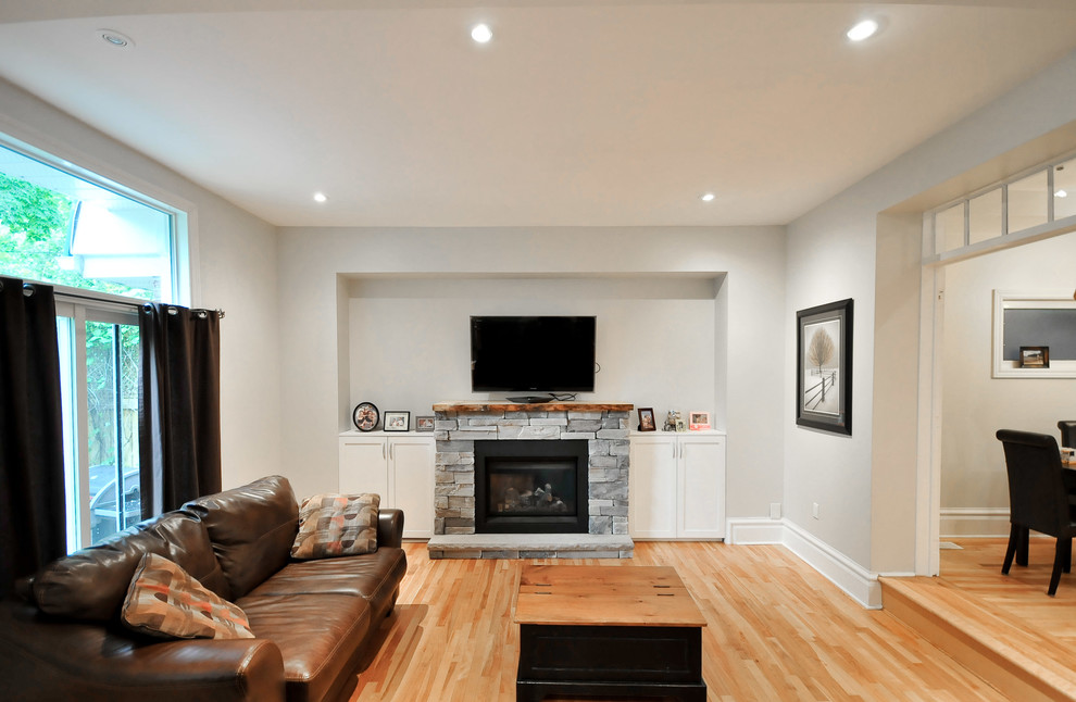 Modelo de sala de estar abierta moderna grande con televisor colgado en la pared, paredes grises, suelo de madera en tonos medios, todas las chimeneas y marco de chimenea de piedra