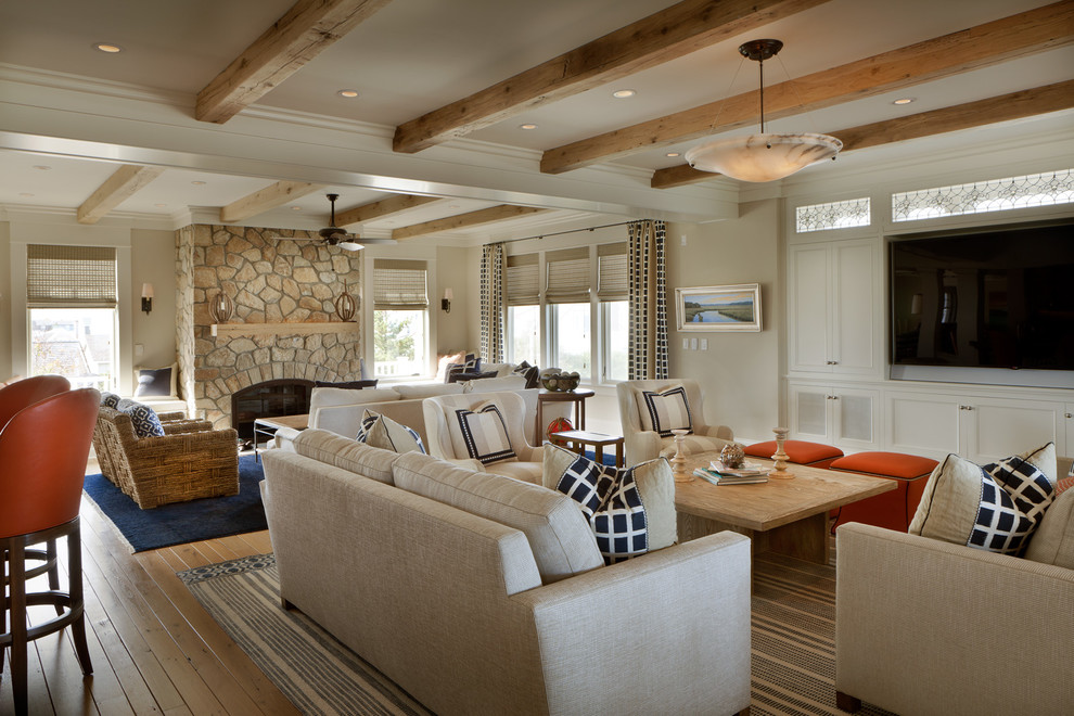 Cette photo montre une grande salle de séjour bord de mer ouverte avec un mur beige, un sol en bois brun, une cheminée standard, un manteau de cheminée en pierre, un téléviseur encastré et un sol marron.