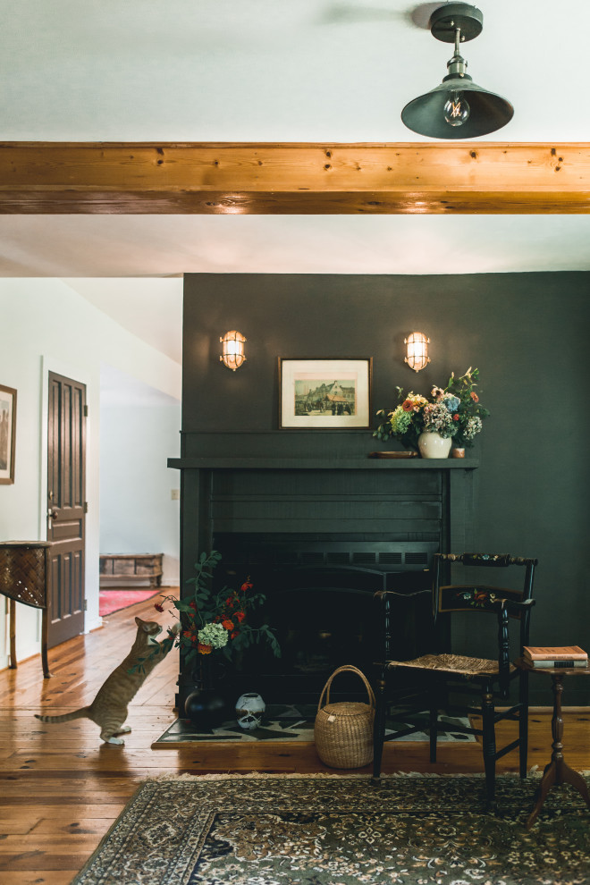 Cette image montre une petite salle de séjour bohème fermée avec un mur noir, un sol en bois brun, une cheminée standard, un manteau de cheminée en bois, un téléviseur d'angle, un sol marron et poutres apparentes.