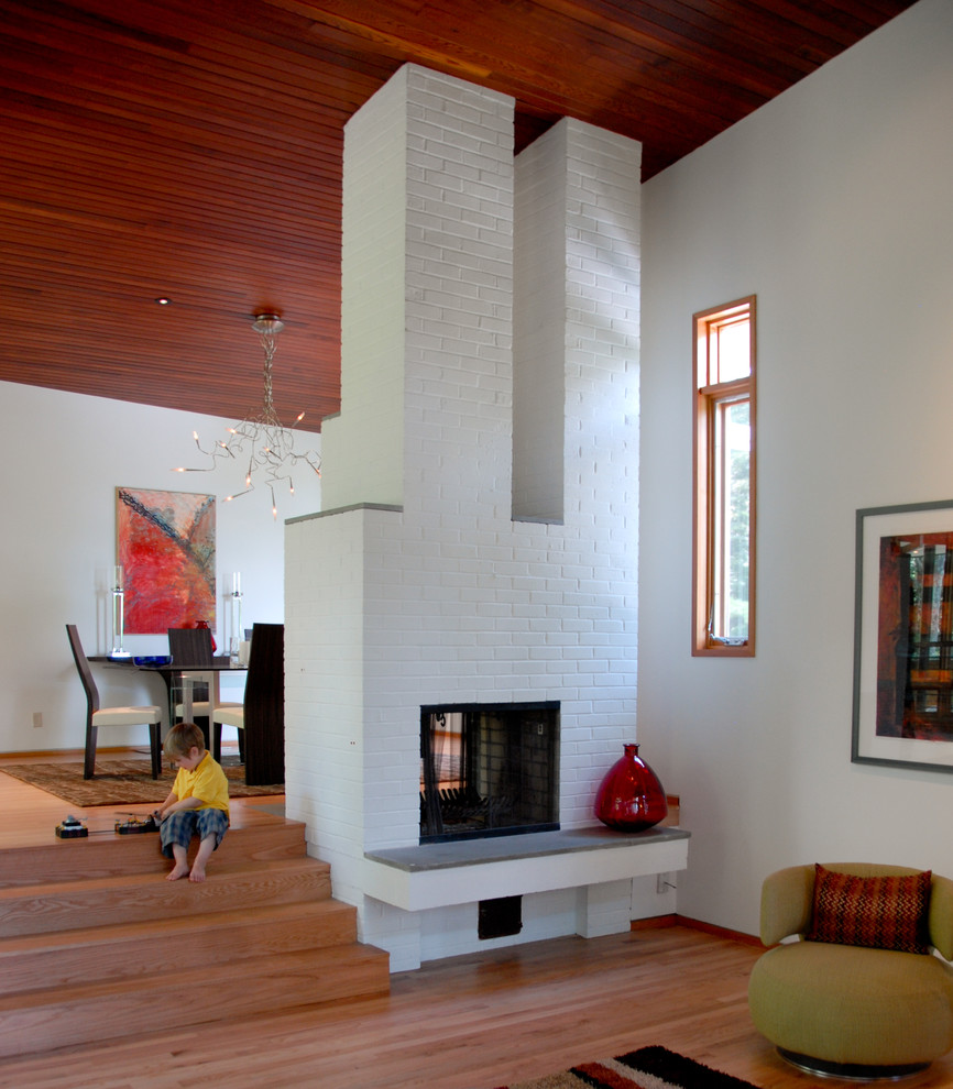 На фото: открытая гостиная комната в современном стиле с белыми стенами, двусторонним камином и фасадом камина из кирпича