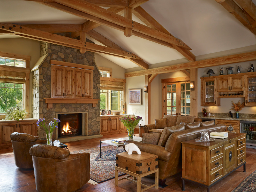 Cette photo montre une grande salle de séjour chic avec un mur beige, un sol en bois brun, une cheminée standard, un manteau de cheminée en pierre, un téléviseur dissimulé et un bar de salon.
