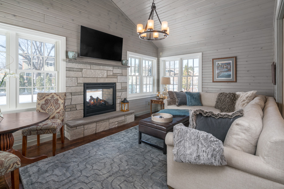 Imagen de sala de estar tradicional renovada con paredes grises, suelo de madera oscura, chimenea de doble cara, marco de chimenea de piedra, televisor independiente y suelo marrón