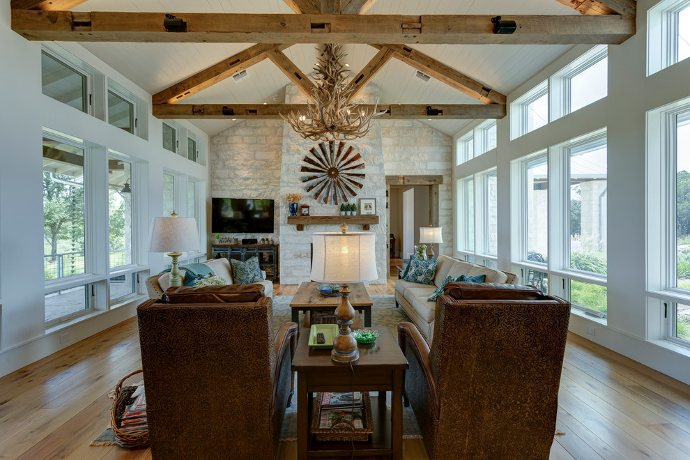 Cette photo montre une grande salle de séjour nature avec un mur blanc, un sol en bois brun, une cheminée standard, un manteau de cheminée en pierre, un téléviseur fixé au mur et un sol marron.