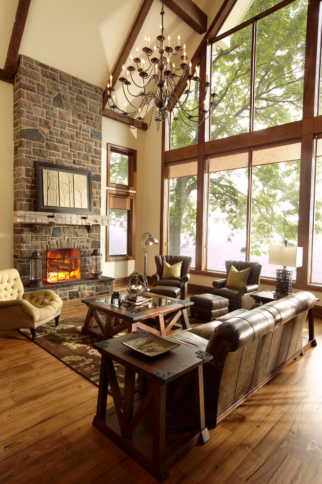 Cette image montre un grand salon chalet avec un manteau de cheminée en pierre, un mur beige, un sol en bois brun, une cheminée standard et éclairage.