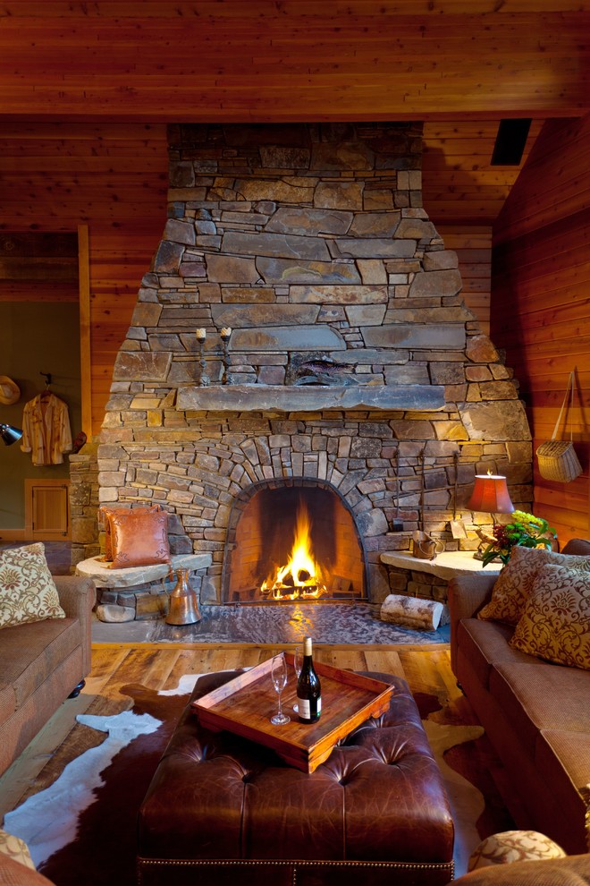 Cette photo montre une grande salle de séjour montagne ouverte avec un mur marron, un sol en bois brun, une cheminée standard, un manteau de cheminée en pierre et aucun téléviseur.