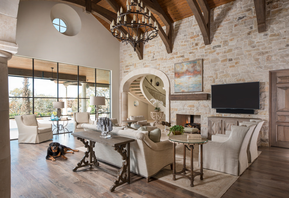 This is an example of an expansive mediterranean open plan games room in Dallas with a standard fireplace, a stone fireplace surround, a wall mounted tv, brown floors, medium hardwood flooring and beige walls.