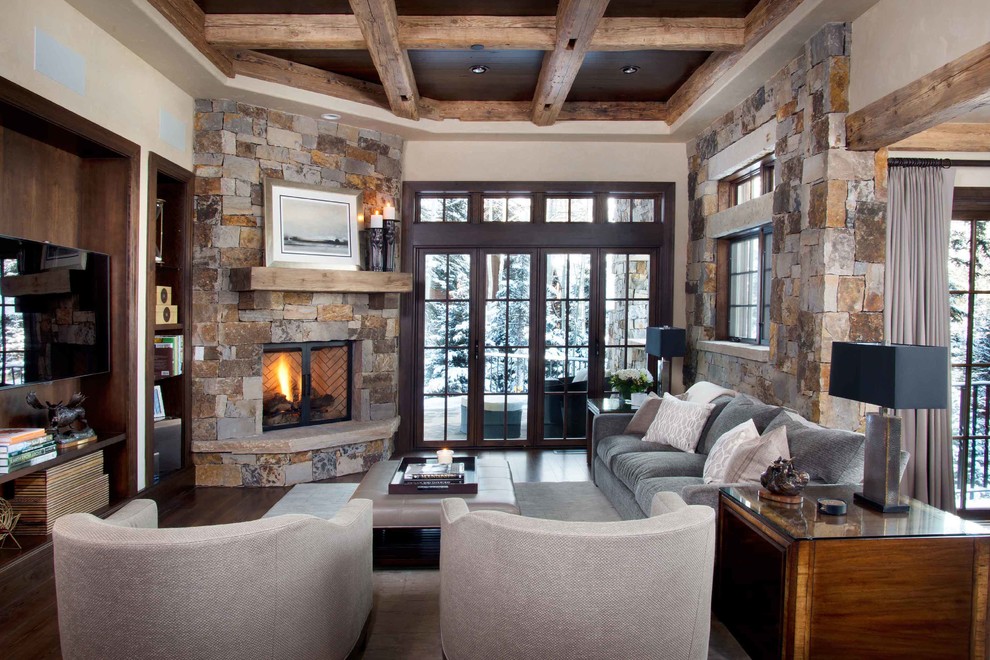 Modelo de sala de estar rústica con paredes beige, chimenea de esquina, marco de chimenea de piedra, televisor colgado en la pared y alfombra