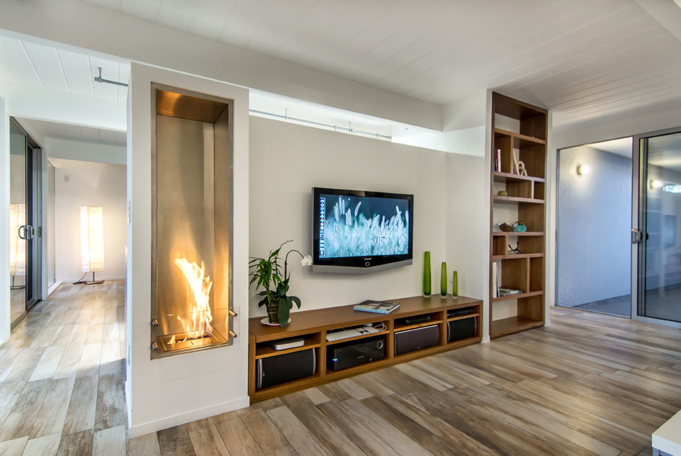 На фото: открытая гостиная комната среднего размера в стиле ретро с белыми стенами, фасадом камина из металла, телевизором на стене, горизонтальным камином, полом из керамогранита и бежевым полом с
