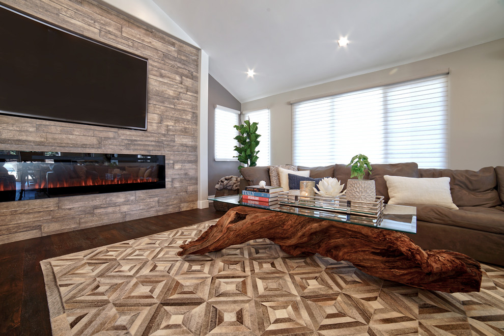 Modelo de sala de estar abierta rural grande con paredes grises, suelo de madera oscura, chimenea lineal, marco de chimenea de piedra, pared multimedia y suelo marrón