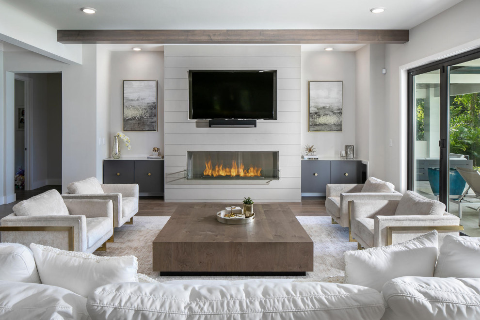 Cette image montre une très grande salle de séjour minimaliste ouverte avec un mur blanc, un sol en bois brun, un manteau de cheminée en lambris de bois, un téléviseur fixé au mur, un sol marron et poutres apparentes.