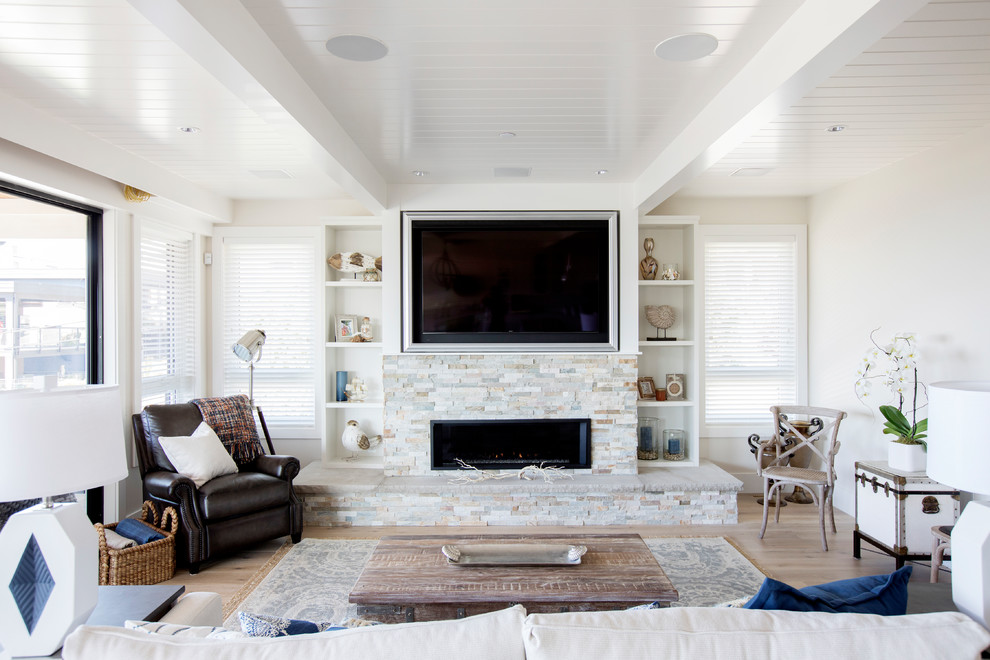 Imagen de sala de estar cerrada costera de tamaño medio con paredes blancas, suelo de madera clara, marco de chimenea de piedra, televisor colgado en la pared, chimenea lineal y suelo beige