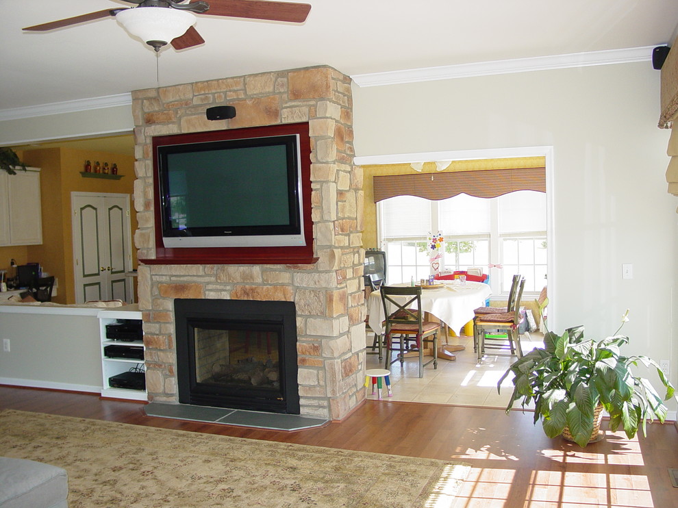 Modelo de sala de estar abierta tradicional grande con suelo de madera en tonos medios, chimenea de doble cara, marco de chimenea de piedra y televisor colgado en la pared