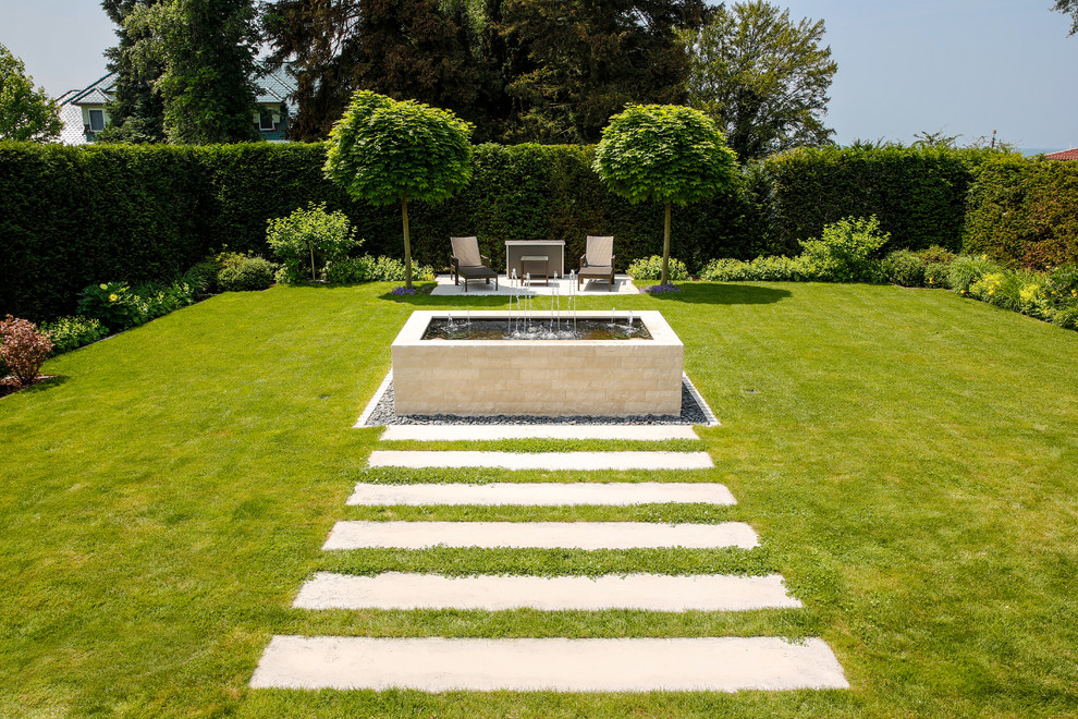 Modelo de jardín actual grande en verano en patio trasero con fuente, adoquines de piedra natural, jardín francés y exposición total al sol