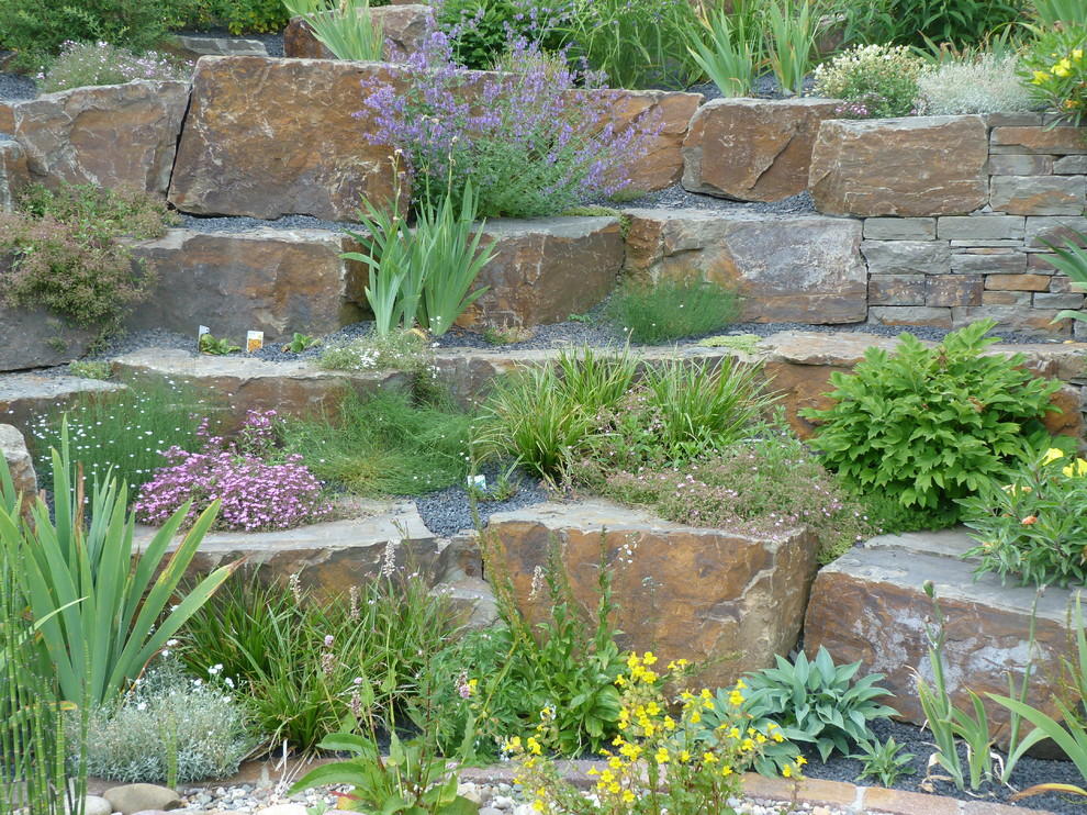 Diseño de jardín campestre grande en verano en ladera con muro de contención, exposición total al sol y adoquines de piedra natural