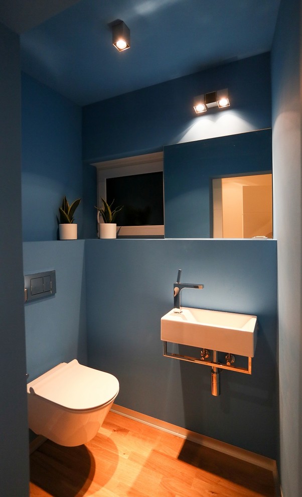 Immagine di un piccolo bagno di servizio minimal con ante bianche, WC sospeso, pareti blu, pavimento in legno massello medio, soffitto in carta da parati e carta da parati