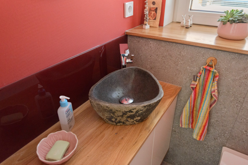 Foto på ett litet funkis brun toalett, med bruna skåp, en vägghängd toalettstol, röd kakel, glaskakel, röda väggar, vinylgolv, ett fristående handfat, träbänkskiva och grått golv