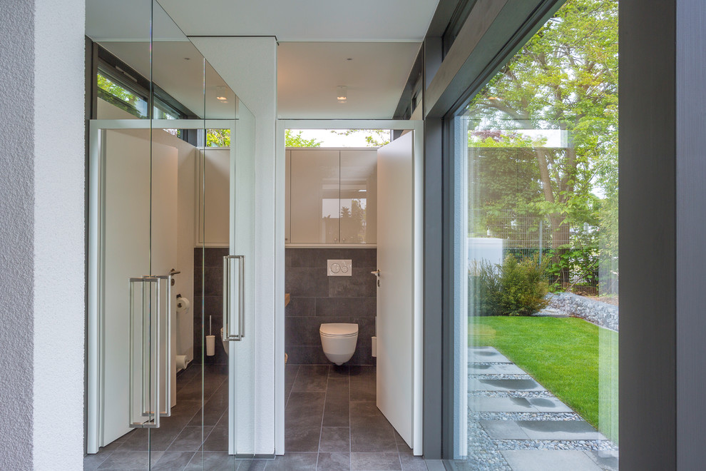 Photo of a large contemporary cloakroom in Other with glass-front cabinets, white cabinets, a wall mounted toilet, grey tiles, ceramic tiles, white walls, ceramic flooring, wooden worktops, grey floors, beige worktops and a vessel sink.