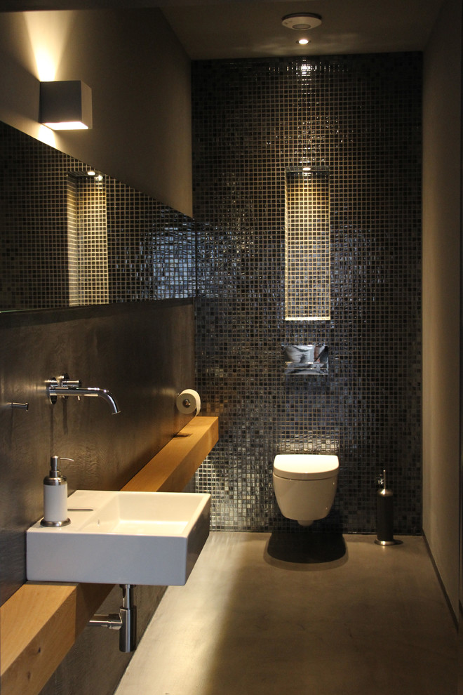 Kleine Moderne Gästetoilette mit Wandtoilette mit Spülkasten, schwarzen Fliesen, Mosaikfliesen, schwarzer Wandfarbe, Betonboden, Wandwaschbecken, Waschtisch aus Holz, grauem Boden und brauner Waschtischplatte in Köln
