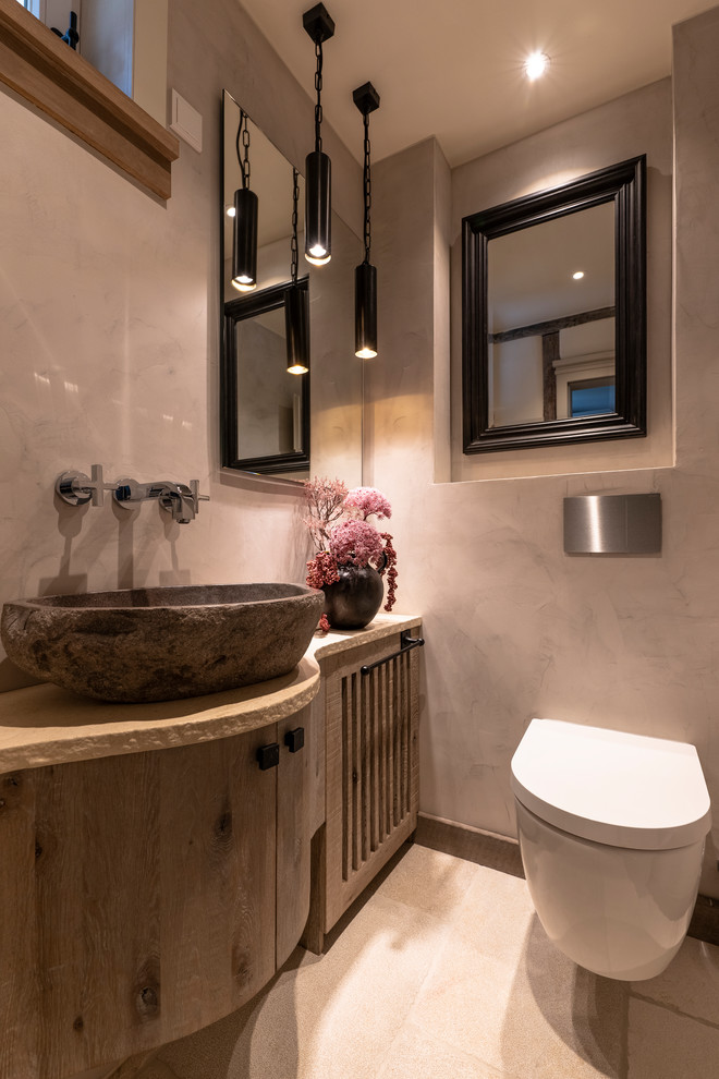 Inspiration for a small country cloakroom in Frankfurt with raised-panel cabinets, brown cabinets, a wall mounted toilet, grey walls, limestone flooring, a vessel sink, beige floors, beige worktops, limestone tiles and solid surface worktops.