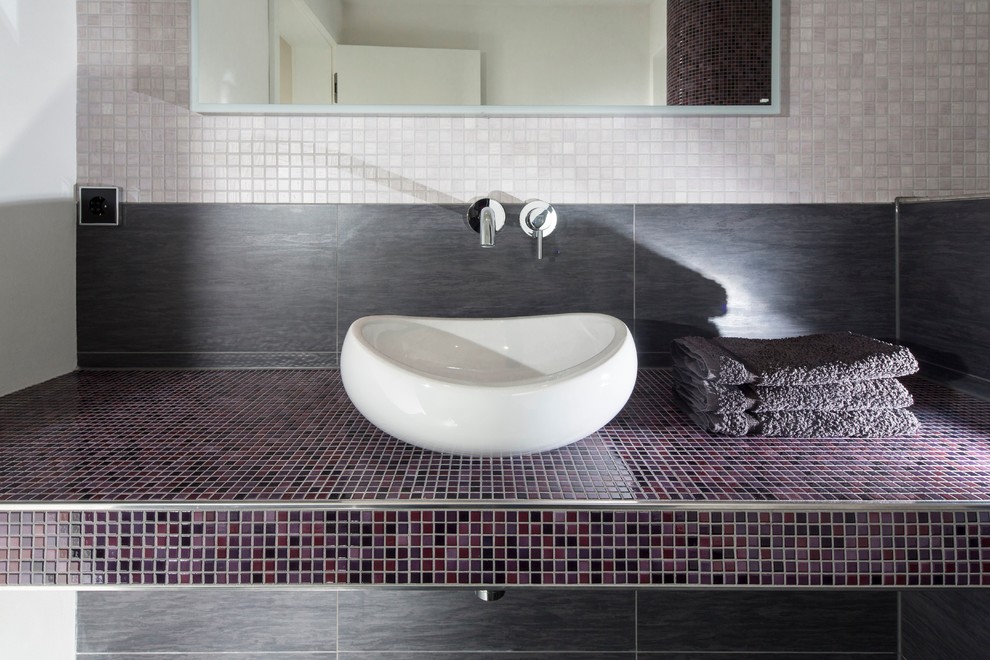 Bild på ett litet funkis lila lila toalett, med ett fristående handfat, kaklad bänkskiva, en toalettstol med separat cisternkåpa, mosaik, vita väggar och flerfärgad kakel