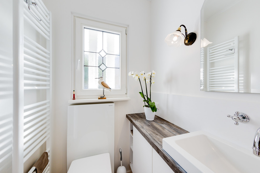 Photo of a small beach style cloakroom in Hamburg with flat-panel cabinets, white cabinets, a wall mounted toilet, white tiles, mirror tiles, white walls, ceramic flooring, a vessel sink, beige floors, wooden worktops and brown worktops.