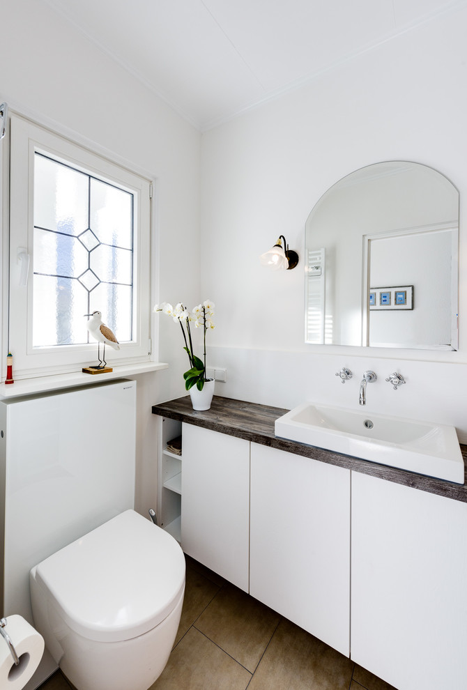 Inspiration for a small contemporary cloakroom in Hamburg with flat-panel cabinets, white cabinets, a wall mounted toilet, white tiles, mirror tiles, white walls, ceramic flooring, a vessel sink, beige floors, wooden worktops and brown worktops.