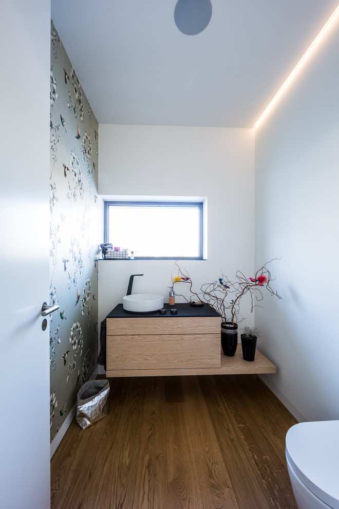 Réalisation d'un WC suspendu design en bois clair de taille moyenne avec un placard à porte plane, un mur multicolore, parquet clair, un sol marron, une vasque et un plan de toilette noir.