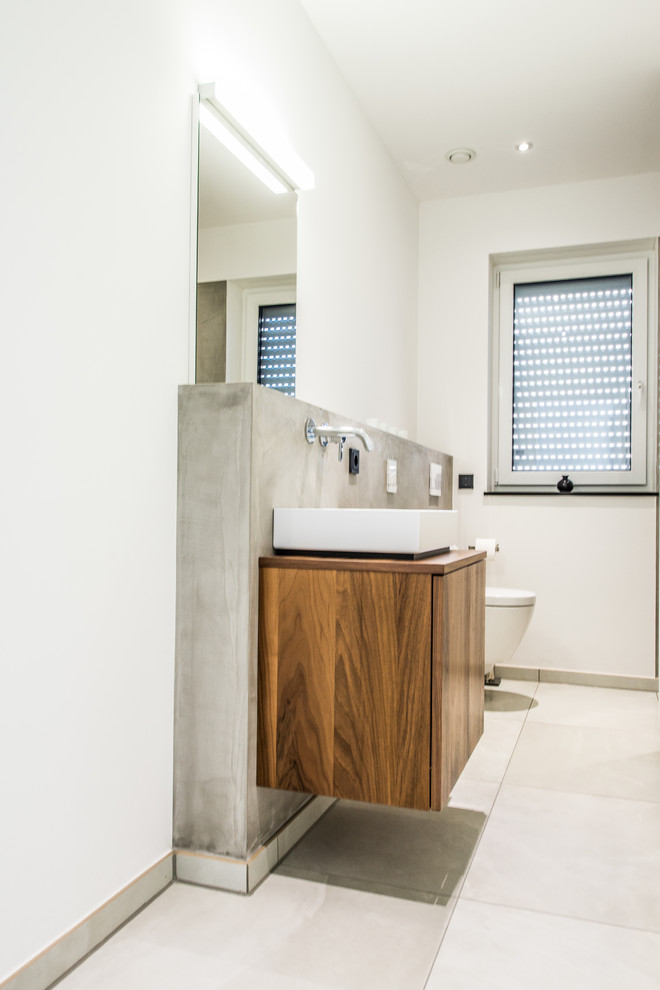 This is an example of a medium sized contemporary cloakroom in Dusseldorf with flat-panel cabinets, dark wood cabinets, a wall mounted toilet, grey tiles, ceramic tiles, white walls, concrete flooring, a vessel sink and wooden worktops.