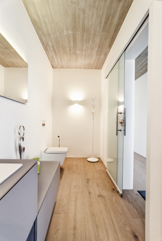 Photo of a large contemporary cloakroom in Other with flat-panel cabinets, grey cabinets, a two-piece toilet, grey walls, light hardwood flooring, an integrated sink and grey worktops.