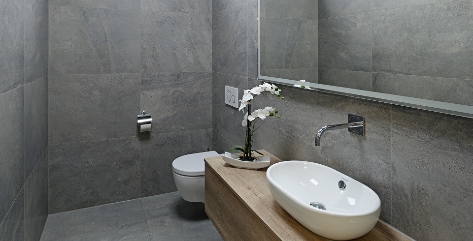 This is an example of a small contemporary cloakroom in Stuttgart with glass-front cabinets, medium wood cabinets, a wall mounted toilet, grey tiles, stone tiles, grey walls, ceramic flooring, a vessel sink, wooden worktops and grey floors.
