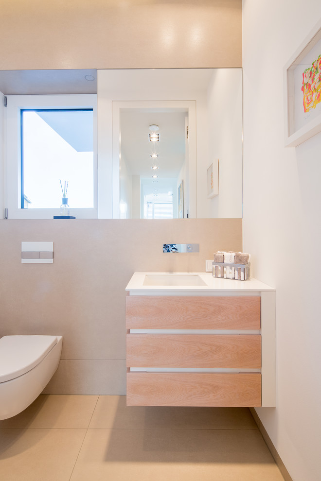 Aménagement d'un petit WC suspendu contemporain en bois clair avec un placard à porte plane, un carrelage beige, un carrelage de pierre, un mur blanc, un lavabo encastré, un plan de toilette en surface solide, un sol beige et un plan de toilette blanc.