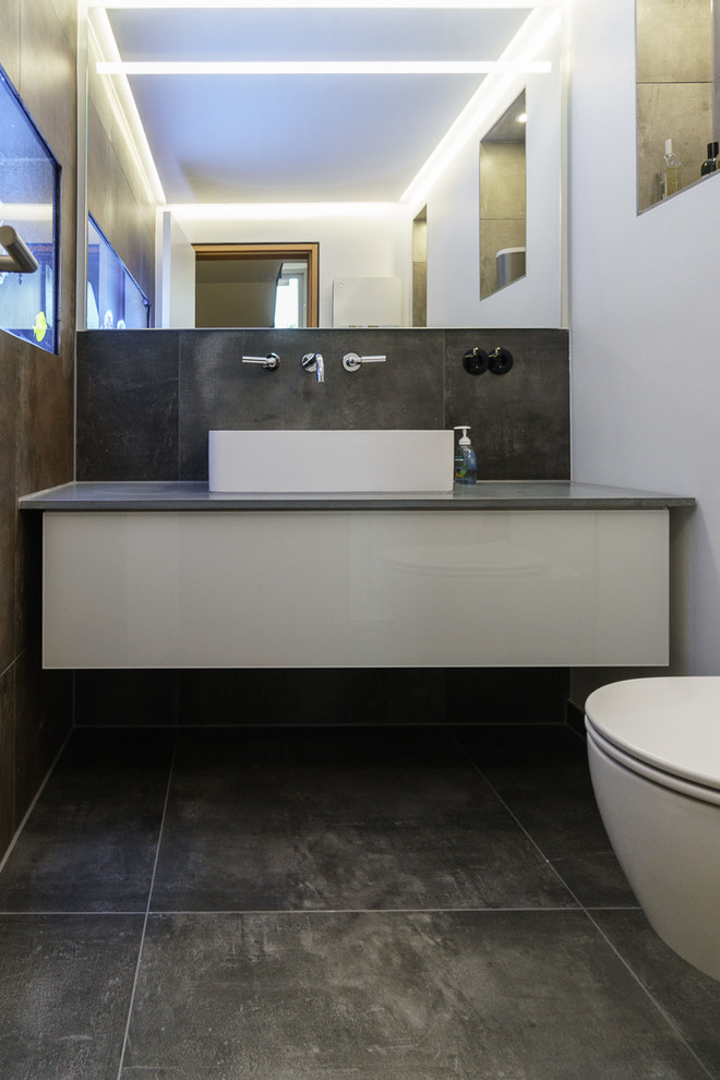 Contemporary cloakroom in Other with flat-panel cabinets, white cabinets, a two-piece toilet, grey tiles, white walls, ceramic flooring and a vessel sink.
