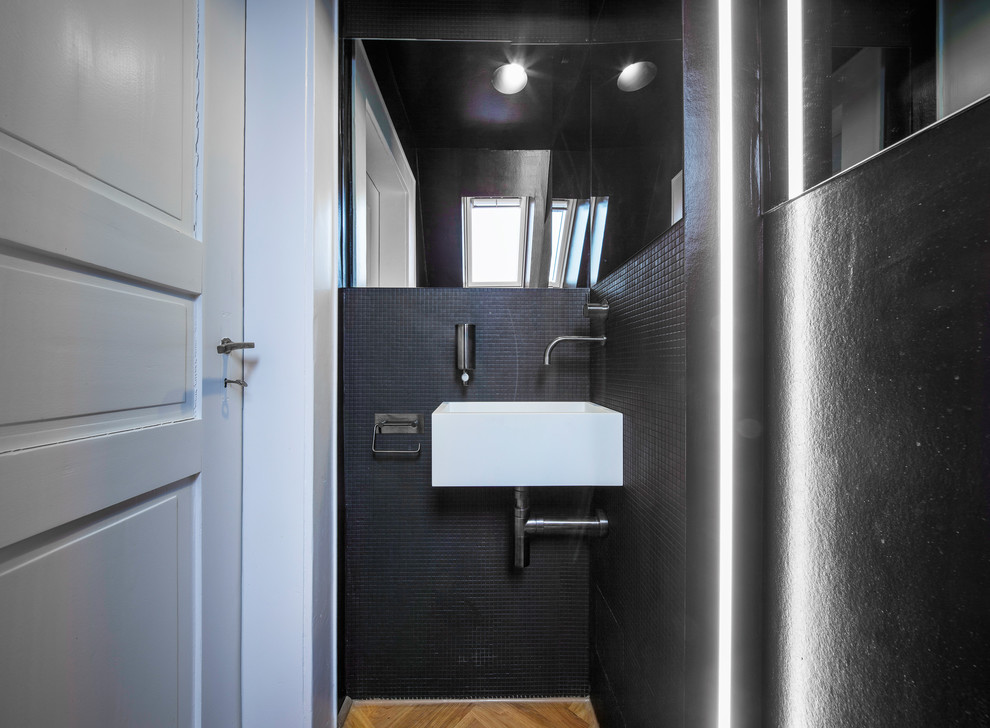 Cette image montre un petit WC et toilettes design avec un carrelage noir, mosaïque, un mur noir, un sol en bois brun, un lavabo suspendu et un sol marron.