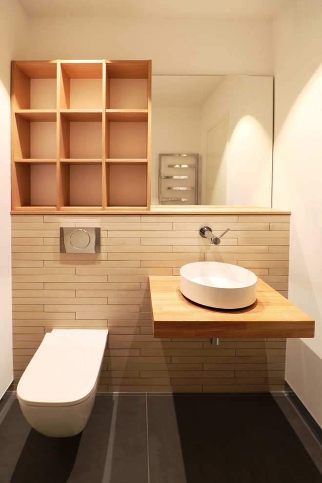 Inspiration för ett litet funkis toalett, med öppna hyllor, skåp i ljust trä, en vägghängd toalettstol, beige kakel, stickkakel, vita väggar, cementgolv, ett fristående handfat, träbänkskiva och brunt golv