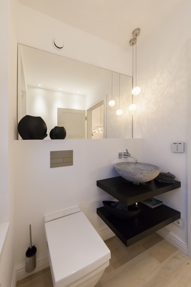 This is an example of a small contemporary cloakroom in Hamburg with open cabinets, black cabinets, a two-piece toilet, white walls, light hardwood flooring, a vessel sink, wooden worktops, beige floors and black worktops.
